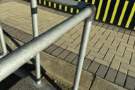 A metal pipe connector on a railing used as a protective barrier or guardrail along a pathway, road, or sidewalk.